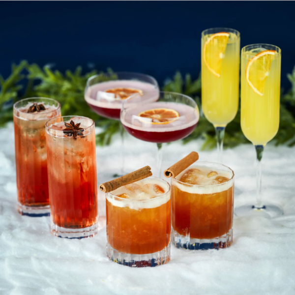 Fem cocktails placeret i sneen; høje glas med stjerneanis, lave glas med kanelstænger, to coupe-glas med tørrede appelsinskiver, og to champagneglas med appelsinskiver. Baggrunden er mørkeblå med grønne grene.