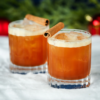 To glas med brun cocktail pyntet med kanelsstang står på en snehvid overflade, omgivet af grønne grene og røde dekorationer, hvilket skaber en festlig atmosfære.