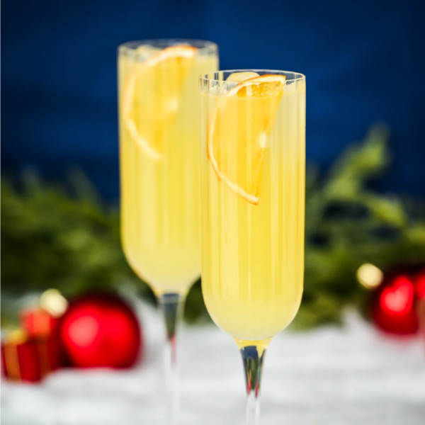 To champagneglas med mimosa cocktails, pyntet med appelsinskiver, står på et bord. I baggrunden er der grønne grene og røde julekugler mod en mørk blå baggrund.