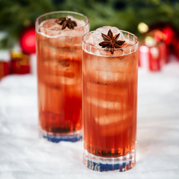 To høje glas med rød væske og isterninger, pyntet med stjerneanis, står på et hvidt bord. I baggrunden ses juleudsmykning med røde kugler.
